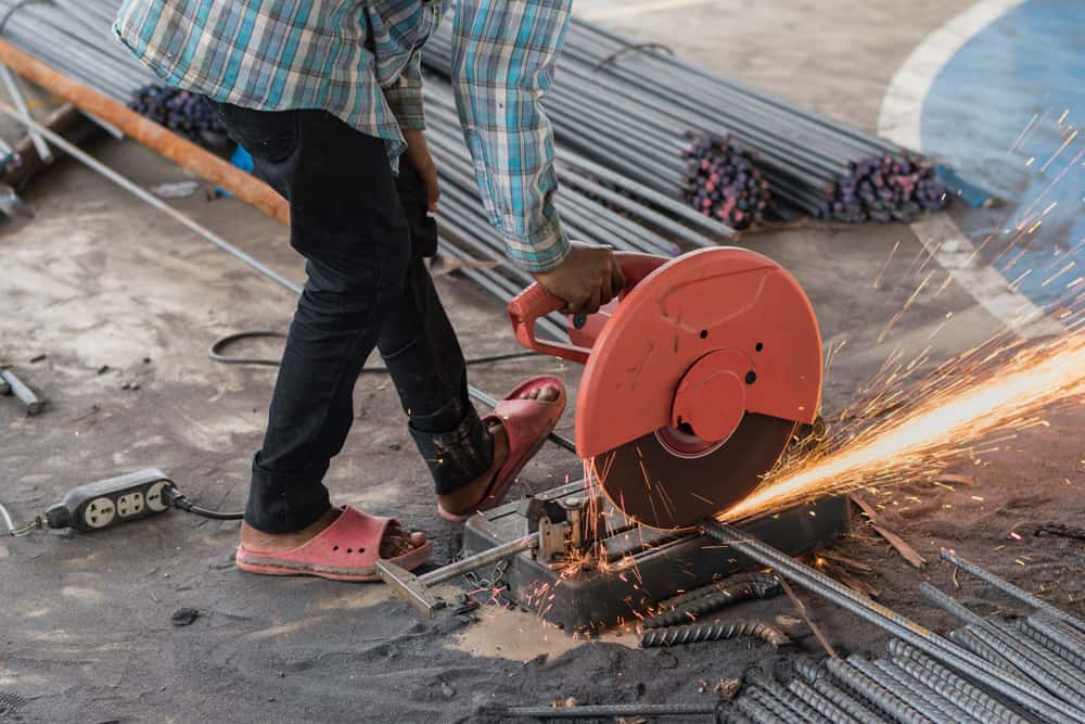 Construction worker cutting rebar with abrasive saw — no eye protection, sandals, sparks flying